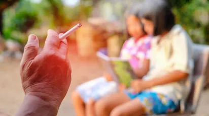 person smoking a cigarette next to children reading a book in a park
