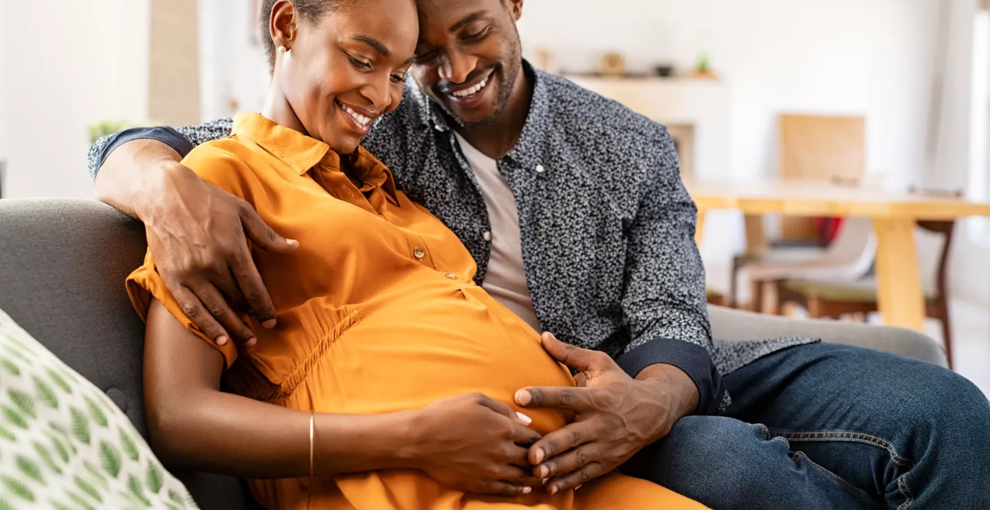 Happy african american husband and pregnant woman hugging belly. Smiling black man hugging happy pregnant wife sitting on sofa and holding tummy. 